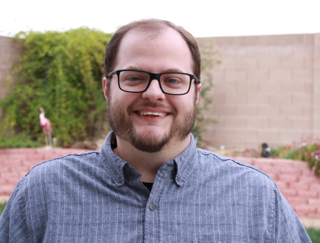 Headshot of Travis Hammond in blue shirt against a brick wall with plants.